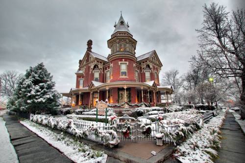 A grand Victorian house covered in snow, surrounded by a snowy garden and trees, under a cloudy sky.