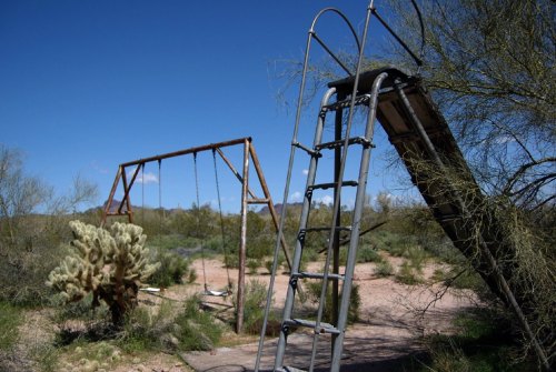 Abandoned playground with swings and a slide, surrounded by desert vegetation under a clear blue sky.