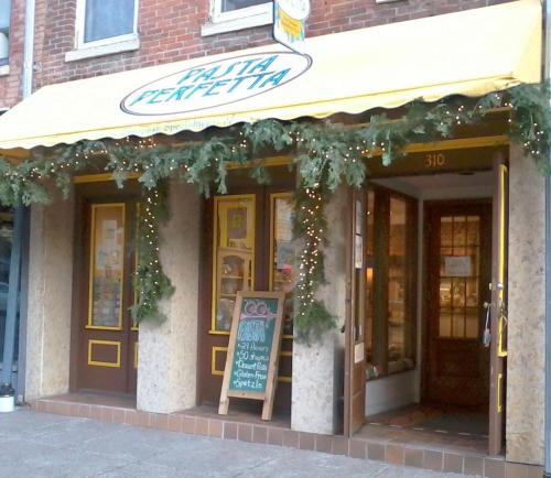 A quaint pasta shop with a yellow awning, festive greenery, and a chalkboard sign outside.