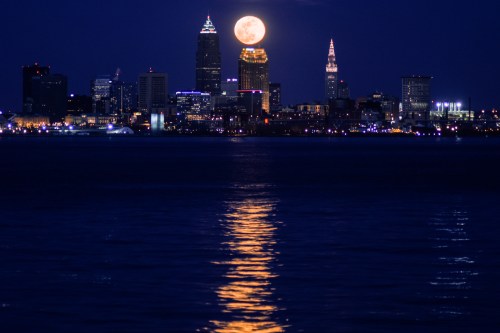 A full moon rises over the Cleveland skyline, reflecting on the water at night. City lights twinkle in the background.