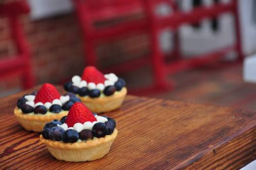 Three fruit tarts topped with strawberries and blueberries, sitting on a wooden table with red chairs in the background.