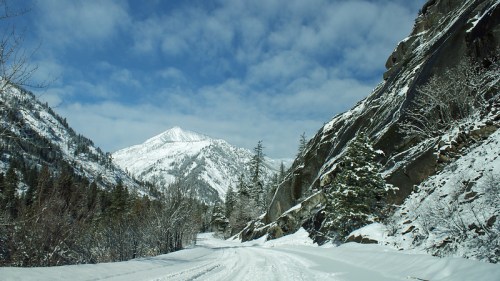 Snow-covered road winding through mountains, flanked by rocky cliffs and trees under a partly cloudy sky.