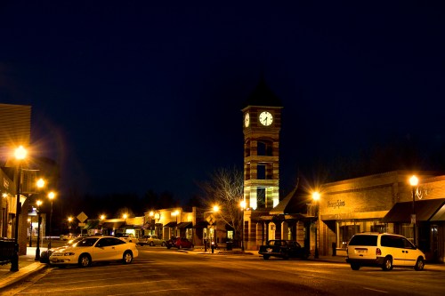 A quiet town square at night, featuring a clock tower and illuminated storefronts along a tree-lined street.