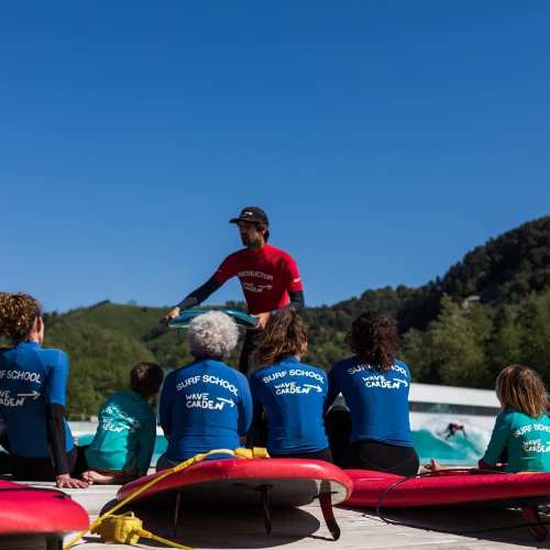 A surf instructor teaches a group of students at a surf school by the water on a sunny day.