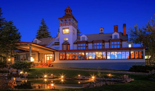 A charming building with a tower, illuminated at dusk, surrounded by greenery and a pond with lights reflecting.