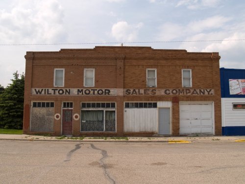 Abandoned brick building with "Wilton Motor Sales Company" painted on the front, featuring boarded windows and a garage door.