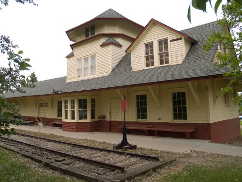 Historic train station with a distinctive tower, featuring a yellow exterior and wooden benches along the platform.