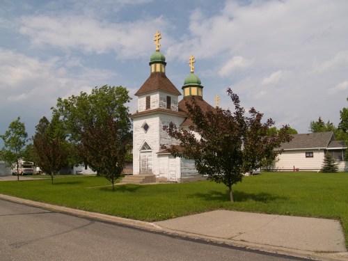 A white church with green domes and golden crosses, surrounded by trees and grass, beside a road under a cloudy sky.