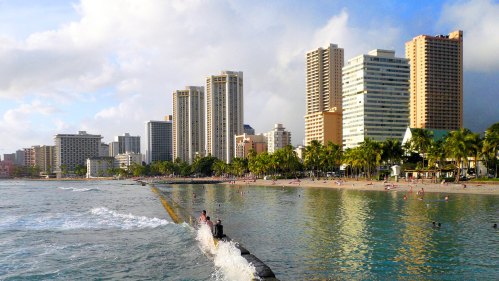 A scenic view of a beach with high-rise buildings, palm trees, and gentle waves under a partly cloudy sky.