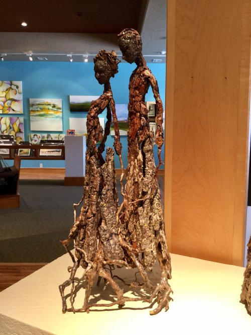 A bronze sculpture of two figures standing closely together, with textured surfaces and elongated forms, displayed indoors.