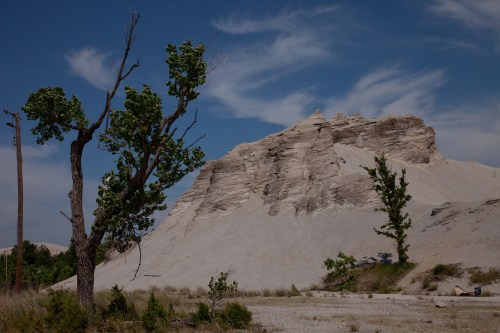 A sandy hill rises against a blue sky, with sparse trees on either side, showcasing a dry, rugged landscape.