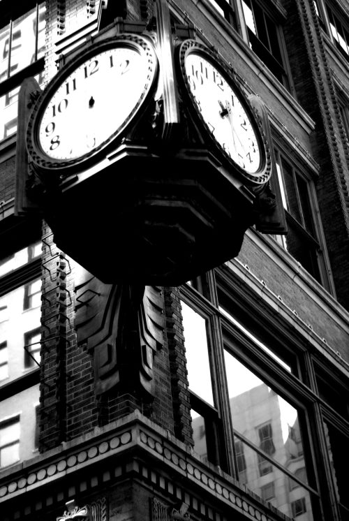 A vintage clock hangs from a brick building, displaying the time in black and white. Reflections are visible in the windows.