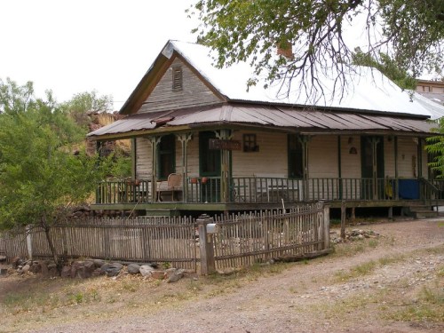A weathered, rustic house with a metal roof, surrounded by a wooden fence and greenery.
