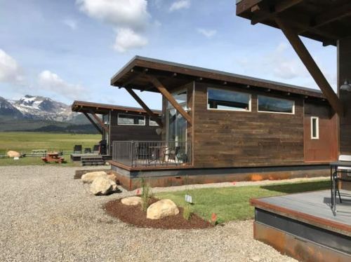 Two modern wooden cabins with large overhangs, set against a backdrop of mountains and a clear blue sky.