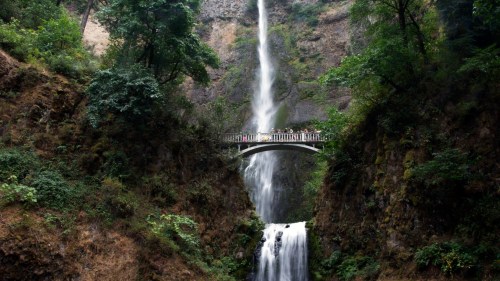 A scenic waterfall cascades down rocky cliffs, with a bridge above surrounded by lush greenery.