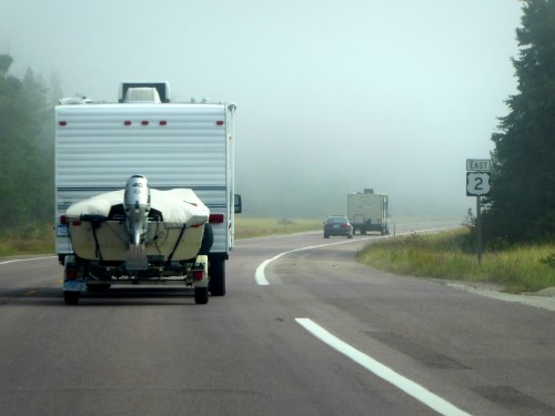 A foggy road scene featuring an RV towing a boat, with another vehicle ahead and trees lining the roadside.