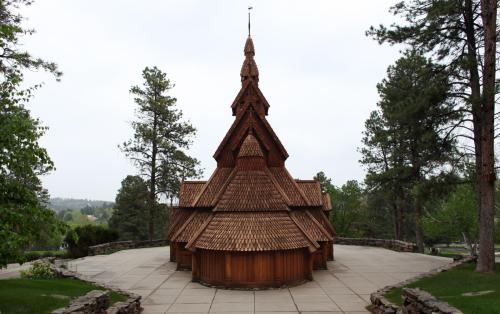 A wooden church with a steeply pitched roof, surrounded by trees and a stone pathway, under a cloudy sky.