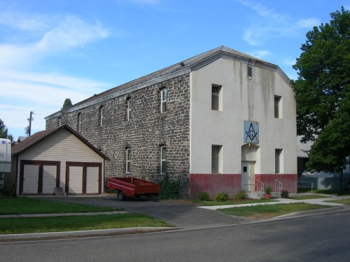 A two-story stone building with a blue Masonic symbol, surrounded by greenery and a red truck parked nearby.
