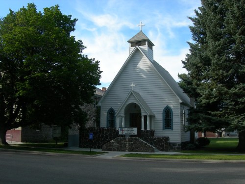 A small white church with a steeple, surrounded by trees and grass, under a blue sky.