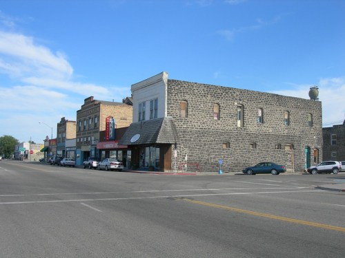 A street view of a small town featuring stone buildings, shops, and parked cars under a blue sky.