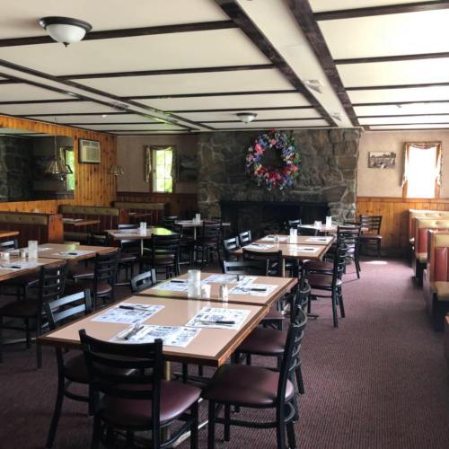 Cozy restaurant interior with wooden walls, tables set for dining, and a stone fireplace adorned with a floral wreath.