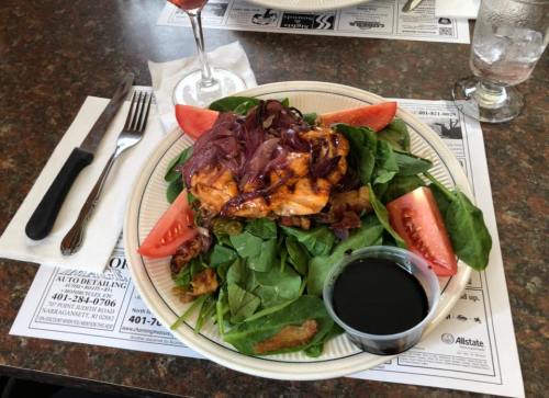 A plate of grilled chicken on a bed of spinach, topped with red onions and tomatoes, served with a side of dressing.