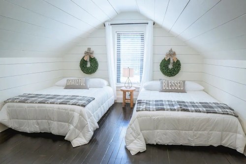 Cozy attic bedroom with two beds, white bedding, and decorative wreaths on the wall, featuring "Grateful" and "Blessed" pillows.