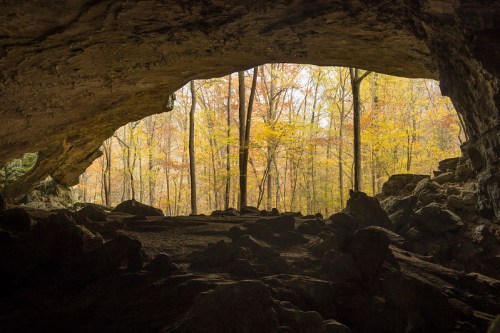 A cave entrance framed by rocks, revealing a forest of autumn trees in the background.