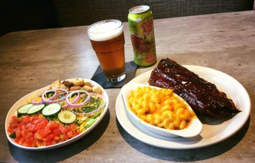 A plate with ribs, mac and cheese, and a salad, alongside a glass of beer and a can of soda on a table.