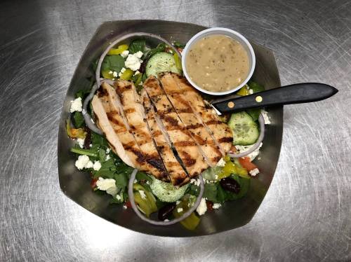 Grilled chicken salad with mixed greens, cucumbers, feta, and a side of dressing in a bowl.