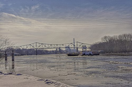 A tugboat navigates a frozen river with a bridge and industrial buildings in the background under a cloudy sky.