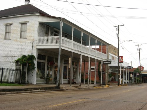 Historic buildings line a quiet street, featuring balconies and storefronts under a cloudy sky. Power lines overhead.
