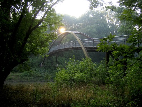 A curved bridge arches over a serene landscape, surrounded by lush greenery and soft morning light.