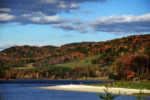 A serene lake surrounded by colorful autumn foliage and a sandy beach under a partly cloudy sky.