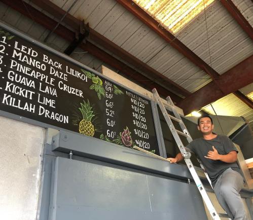 A young man gives a shaka sign next to a chalkboard menu featuring various drinks and prices. A ladder is nearby.