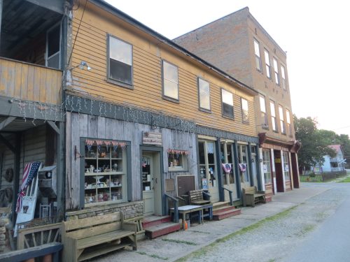 Historic buildings with wooden facades, storefronts, and benches line a quiet street, adorned with patriotic decorations.