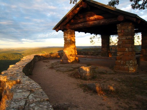 A stone pavilion with a wooden roof overlooks a scenic landscape at sunset, surrounded by rolling hills and trees.
