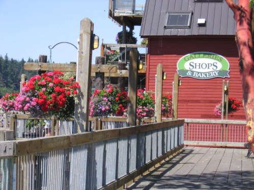 A wooden walkway lined with colorful flower baskets leads to a red building with a sign for galleries, shops, and a bakery.