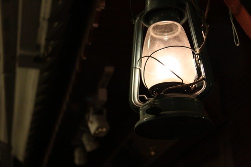A vintage lantern glowing softly in a dimly lit setting, hanging from a wooden structure.