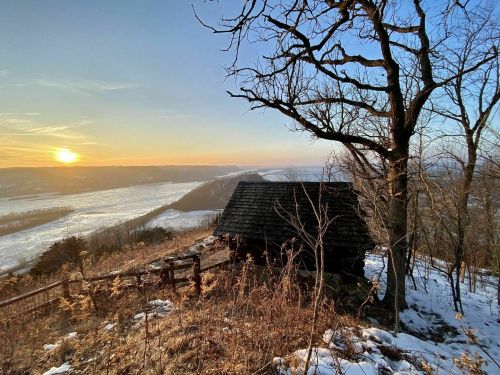 A rustic cabin on a hillside at sunset, surrounded by trees and snow, overlooking a valley.