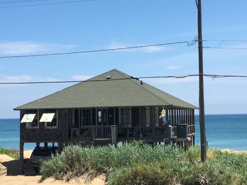 A beach house with a green roof, surrounded by sand and grass, overlooking the ocean under a clear blue sky.