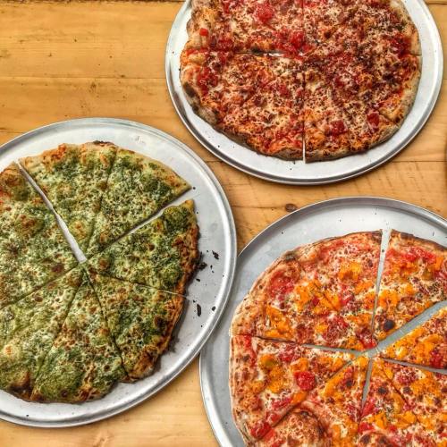 Three pizzas on metal trays: one with green pesto, one with tomato and cheese, and one with orange cheese and toppings.