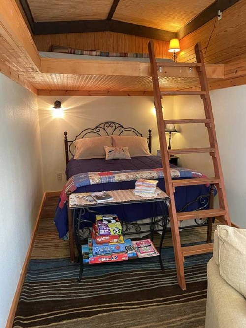 Cozy bedroom with a loft, featuring a bed, wooden ladder, and a table with games and books. Warm lighting enhances the space.