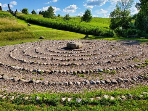 A stone labyrinth with a central rock, surrounded by grass and trees under a blue sky with fluffy clouds.