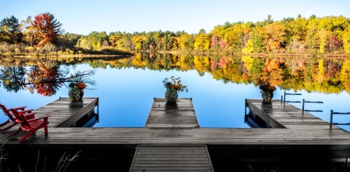 A serene lake view with colorful autumn trees reflecting on the water, featuring a wooden dock and red chairs.