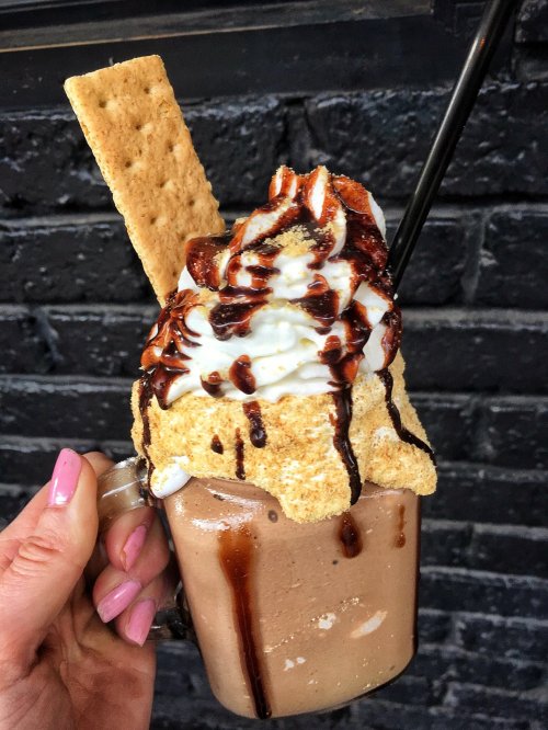 A hand holding a tall milkshake topped with whipped cream, chocolate drizzle, and a graham cracker.
