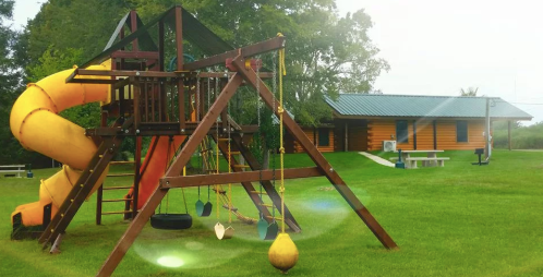 A colorful playground with a yellow slide and swings, set in a grassy area near a log cabin.
