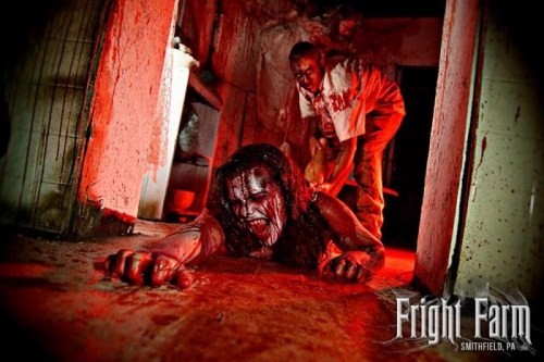 A horror scene with a bloodied woman crawling on the floor while a figure looms behind her in a dimly lit room.