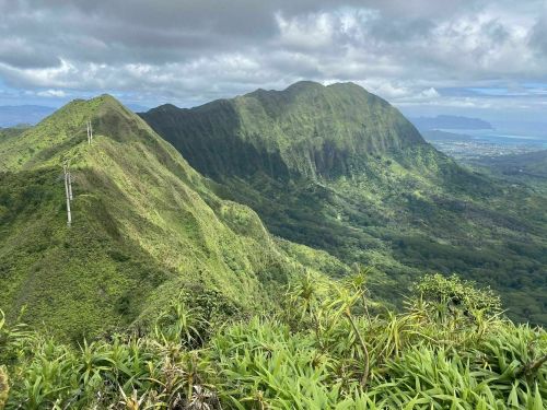 Lush green mountains under a cloudy sky, with a view of the coastline in the distance.