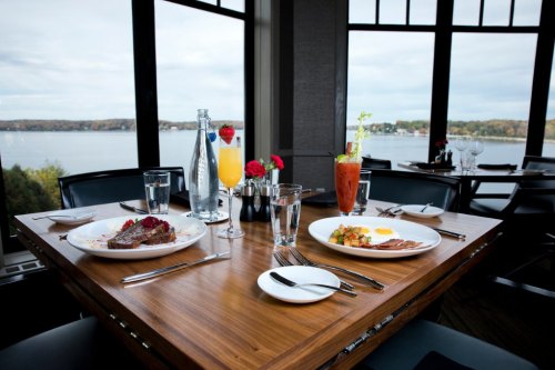 A beautifully set table with two plates of food, drinks, and a scenic view of a lake through large windows.